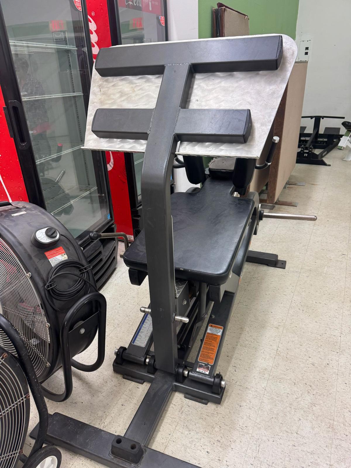 a weight training machine in a gym, with a black and gray color scheme. The machine appears to be a piece of exercise equipment designed for strength training.