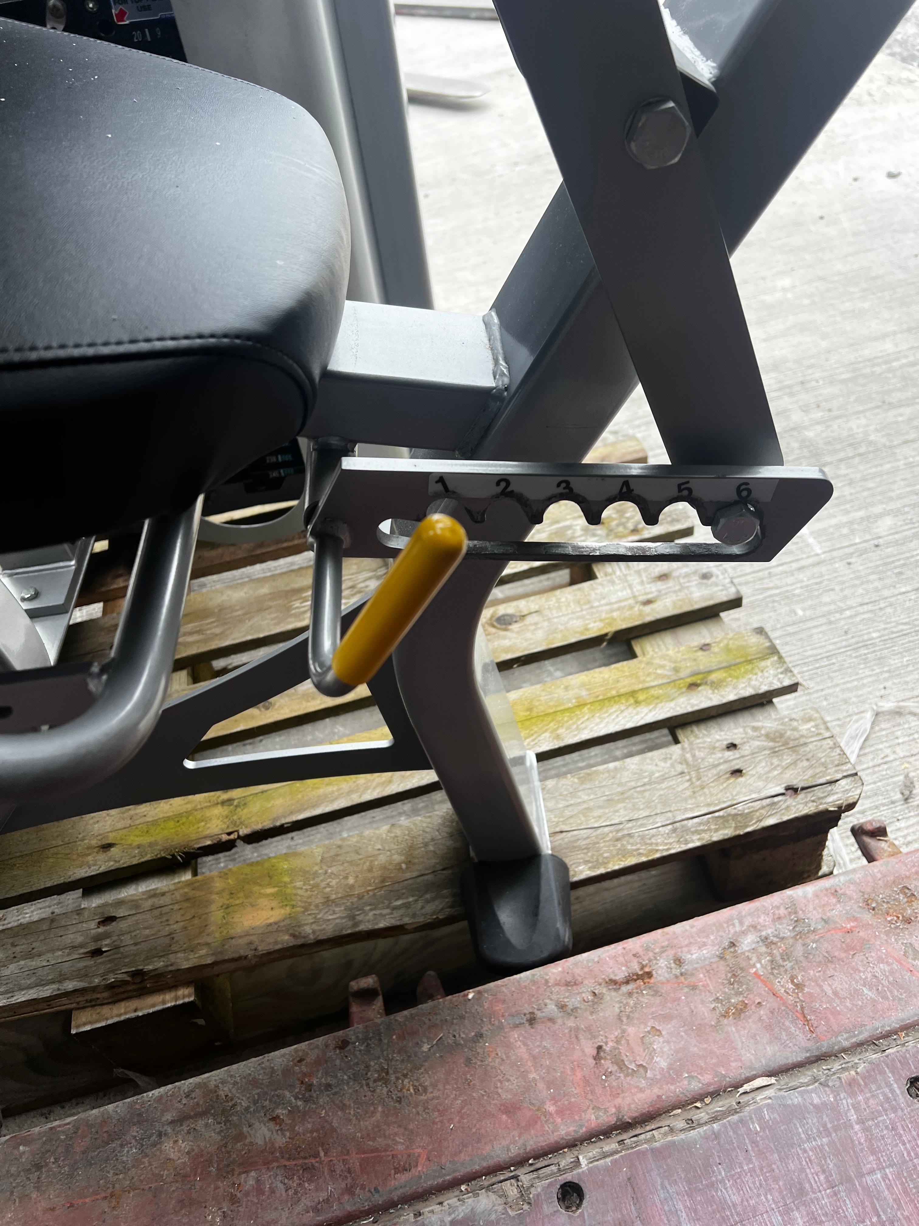a close-up view of a piece of exercise equipment, possibly a weight machine or a bench, with a yellow handle or knob attached to it. The equipment is placed on a wooden pallet or crate, which is positioned on a concrete floor.