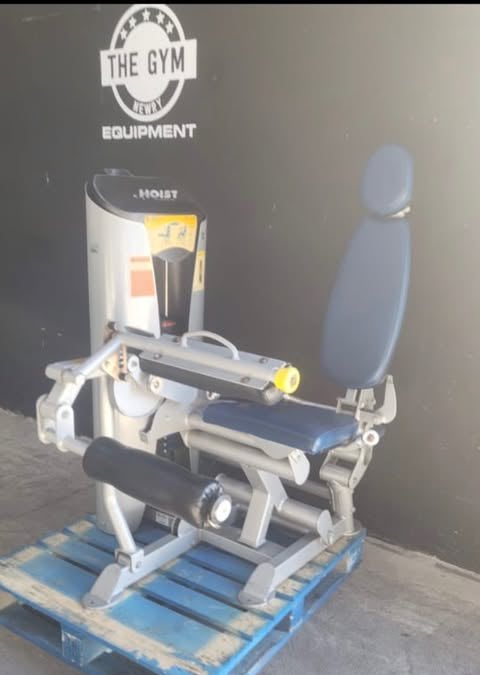 A hoist leg curl machine is displayed on a blue pallet against a black wall with a gym equipment logo.