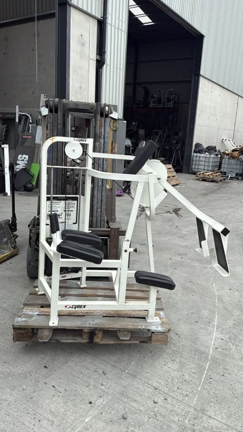 A white and black weight training machine is sitting on a wooden pallet in an outdoor area, with a warehouse or storage building in the background.
