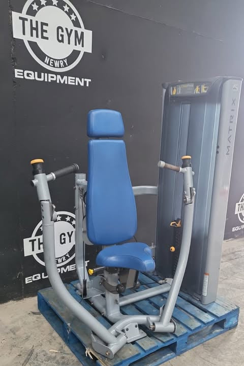 A blue chest press machine is displayed in front of a gym equipment wall, with the text "MATRIX CHEST PRESS" visible.