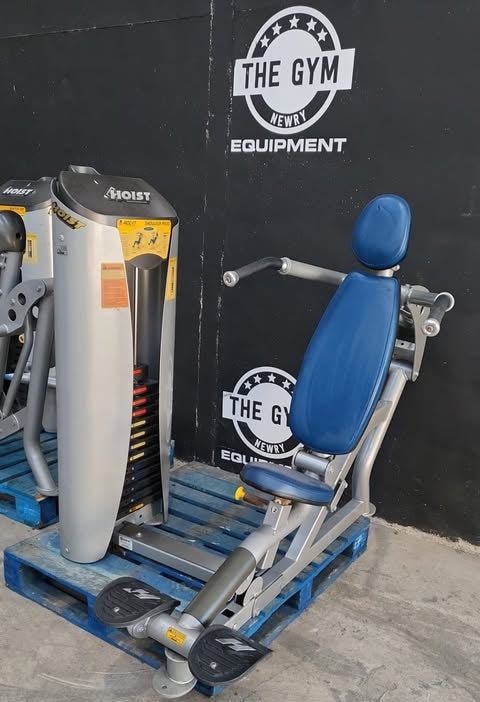 A hoist shoulder press machine is displayed against a black wall with the text "THE GYM NEWR" and "THE GYM EQUIPMENT" visible.