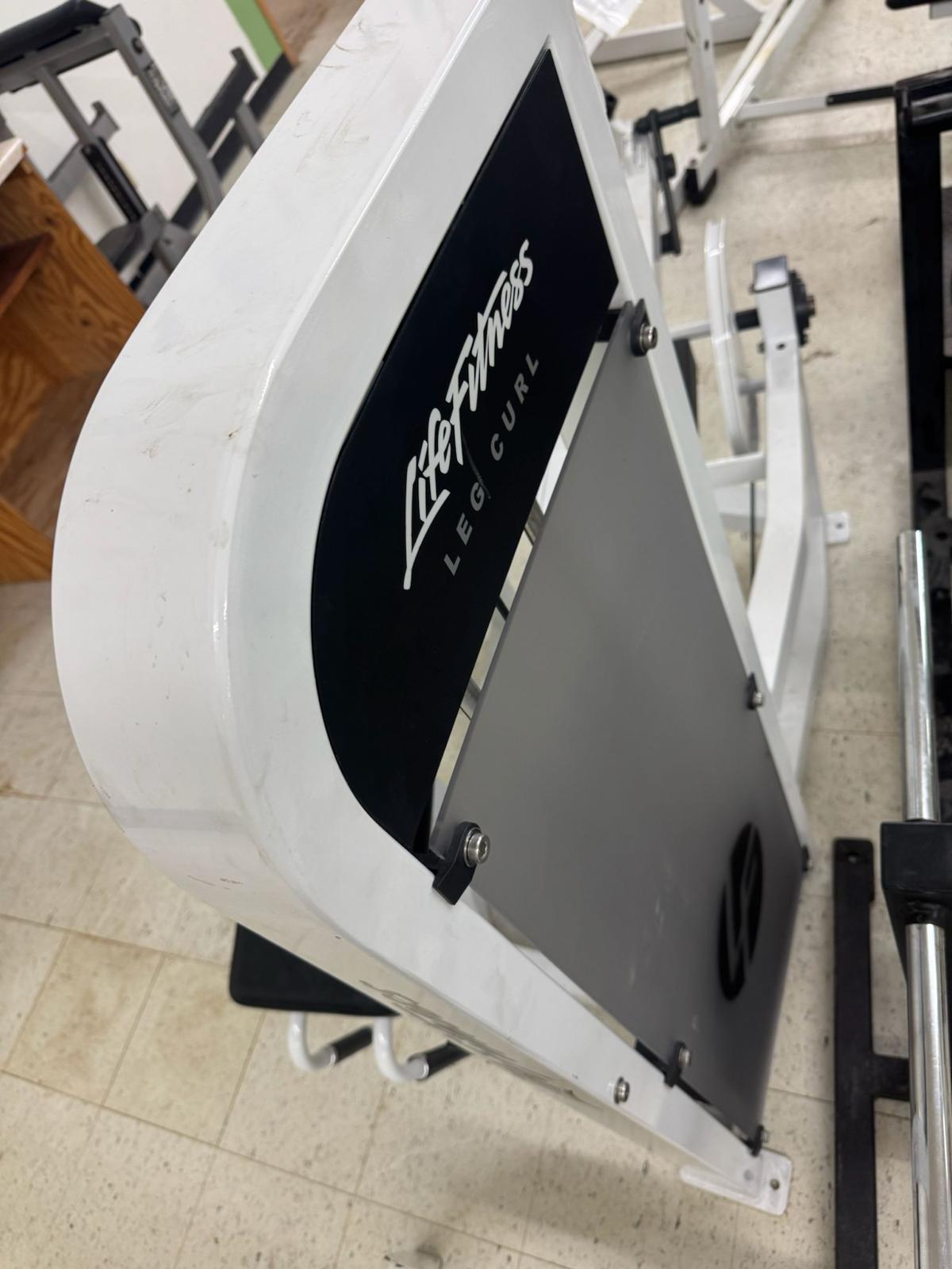 A close-up view of a white and black leg curl machine in a gym, with the brand name "Life Fitness" visible on the machine.