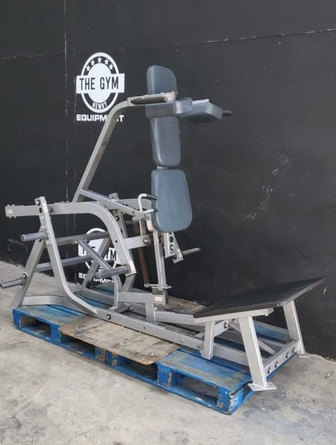 A weight training machine, possibly a leg press or leg curl, is sitting on a blue pallet against a black wall with a logo for "THE GYM" and "equipment" visible.