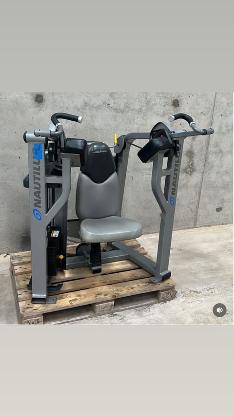 A gray and black weight training machine with a black seat and handles, placed on a wooden pallet against a concrete wall.