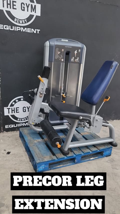 a weight training machine, specifically a precor leg extension, placed on a blue wooden pallet. The machine is positioned in front of a wall with the text "THE GYM NEWRY EQUIPMENT" written on it.