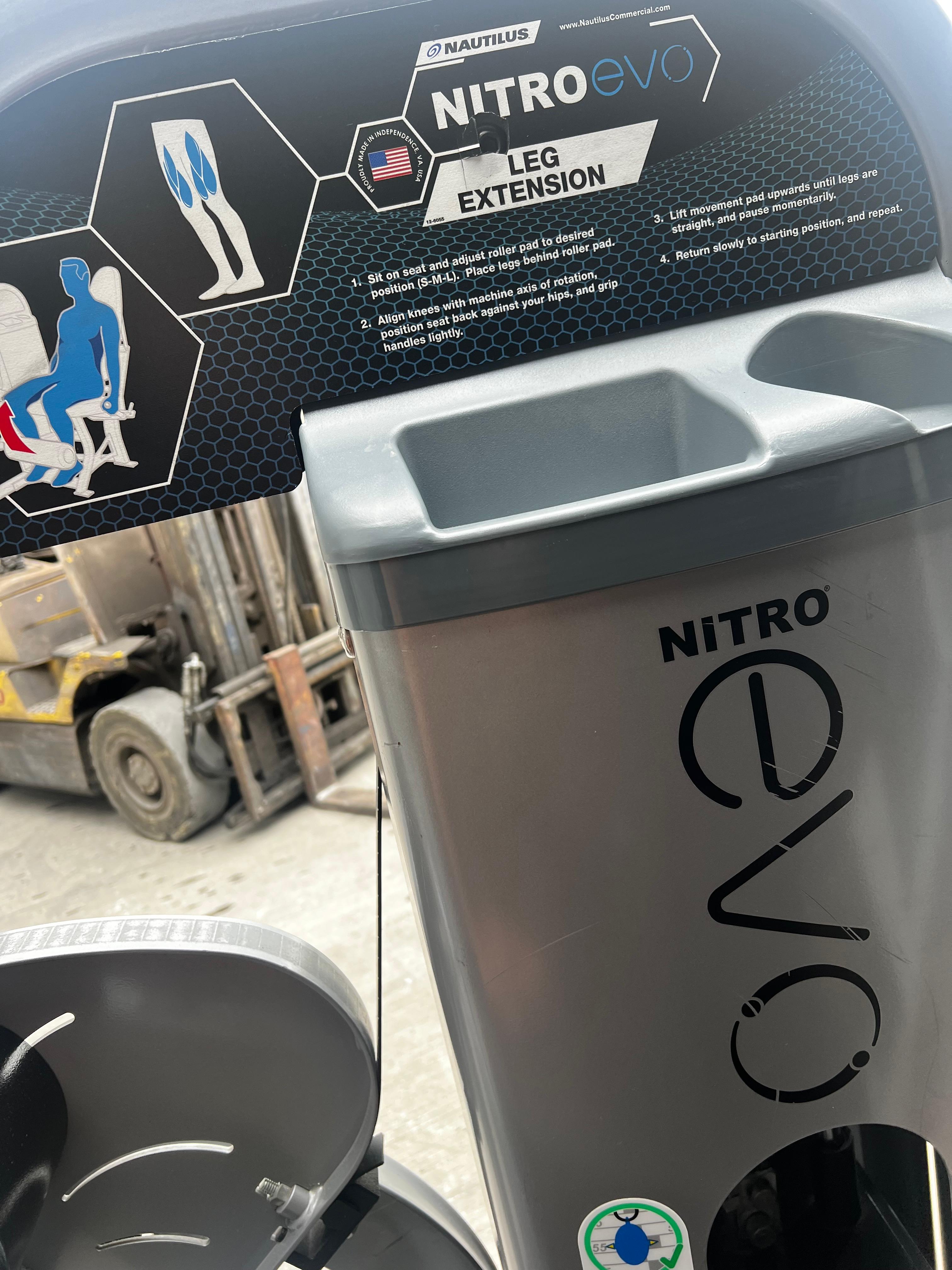 a close-up view of a Nitro Evo leg extension machine, with a focus on the leg extension feature. The machine is placed in an outdoor setting, with a forklift visible in the background.