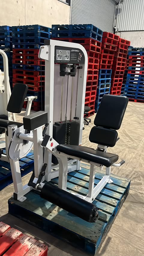 A weight training machine with a black seat and various attachments, situated in a warehouse setting with stacks of pallets in the background.